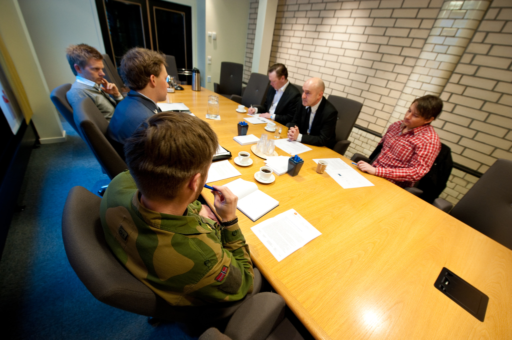 Eine Gruppe von Menschen, die um einen Tisch mit Tassen, B├╝chern, Papieren, einer Flasche, einem Krug und Stiften sitzen.