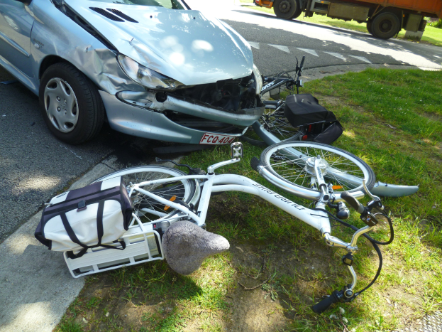Ein verunglücktes Auto am Straßenrand mit einem Fahrrad auf dem Gras, Bäumen und einem klaren blauen Himmel im Hintergrund.