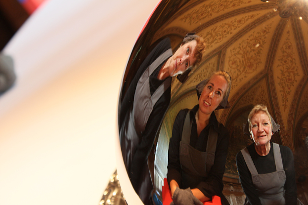 Ein reflektierender Ball auf einem weißen Tisch zeigt das Spiegelbild von drei lächelnden Frauen, die leicht gebückt sind, mit einem Dach im Hintergrund.