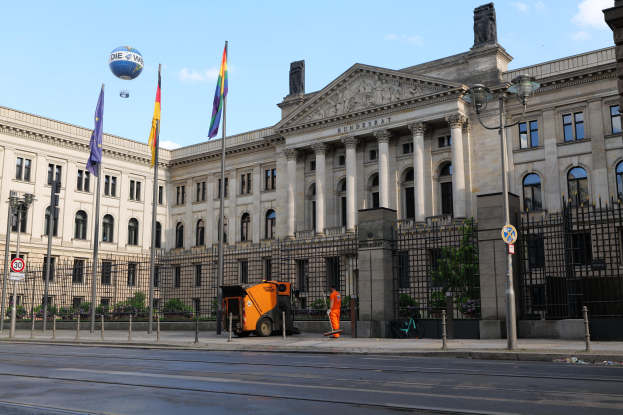 Das Reichstagsgebäude in Berlin, Deutschland, ein großes Gebäude mit zahlreichen Fenstern, umgeben von Laternenmasten, Fahrzeugen, Menschen und Bäumen unter einem bewölkten Himmel.