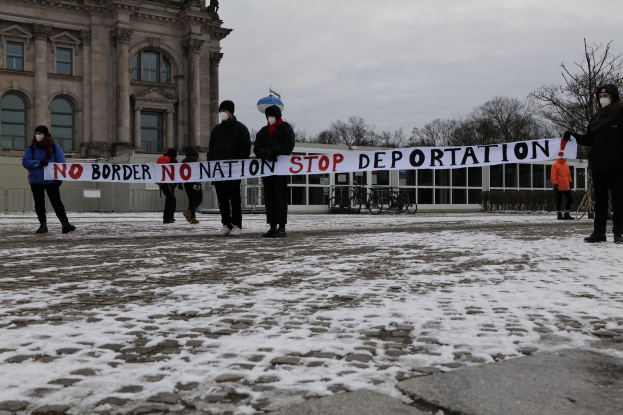 Eine Gruppe von Menschen steht im Schnee und hält ein Banner mit der Aufschrift "Keine Grenze, kein Land, Stopp der Abschiebung", daneben stehen Fahrräder und im Hintergrund sind Bäume und ein klarer blauer Himmel zu sehen.