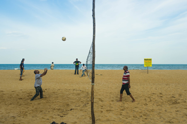 Ein Volleyballnetz mit Spielern auf beiden Seiten, ein Ball in der Luft und Zuschauern sowie ein Warnschild, Wasser und eine bewölkte Himmel im Hintergrund.