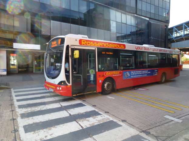 Ein Bus auf einer Straße mit einigen sitzenden Passagieren, einem Gebäude und einer Brücke im Hintergrund.