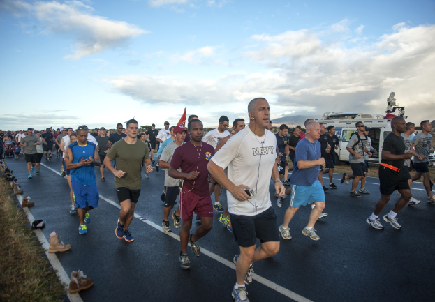 Gruppe von Menschen, die bei einem Marathon auf einer Straße mit Gras, Schuhwerk, Fahrzeugen, Bäumen, Hügeln und einem bewölkten Himmel im Hintergrund laufen.