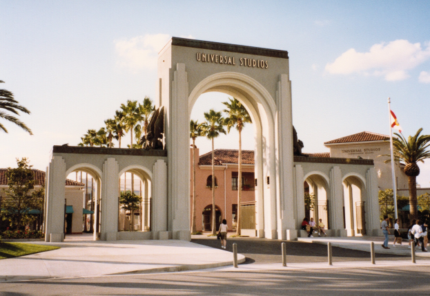 Eingang von Universal Studios Hollywood mit einem Bogen, einer Statue, Menschen, Bäumen, Gebäuden, einer Fahnenstange mit einer Fahne und einem bewölkten Himmel.
