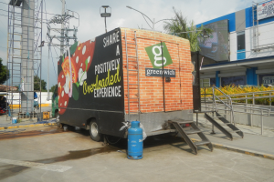 Lastwagen vor einem Gebäude mit einer Tafel, auf der "Teilen Sie ein positiv überladenes Erlebnis" steht, mit Stufen, einem Zylinder auf dem Boden, Geländern, Strommasten mit Drähten, Laternen, Bäumen und einem bewölkten Himmel.