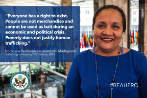 Eine Frau in einem blauen Kleid steht lächelnd vor einem Gebäude mit einem Zitat über Menschenhandel, mit Menschen, Pflanzen und Flaggen im Hintergrund.