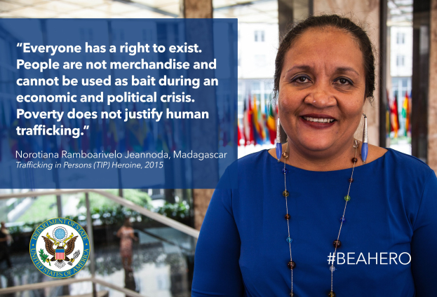 Eine Frau in einem blauen Kleid steht lächelnd vor einem Gebäude mit einem Zitat über Menschenhandel, mit Menschen, Pflanzen und Flaggen im Hintergrund.