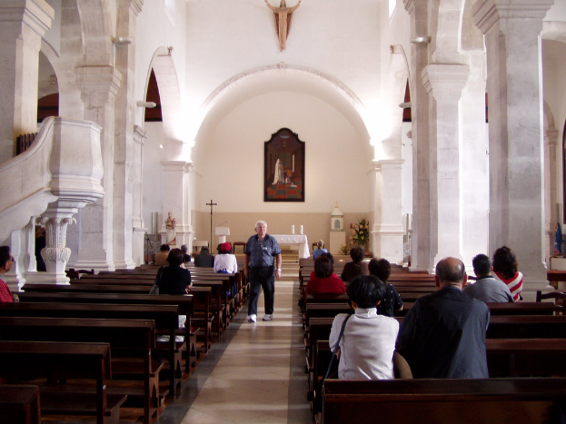 Ein älterer Mann geht den Mittelgang einer Kirche entlang, flankiert von Sitzreihen mit sitzenden Personen, während die Wände im Hintergrund Fotos und Idole zeigen.