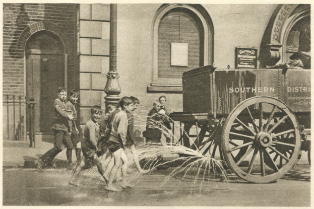 Kinder beim Spielen mit einem Hydranten vor einem Gebäude mit einem Wagen im Hintergrund, schwarz-weiß.