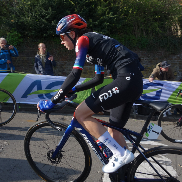 Professionelle Radrennfahrerin Katie Archibald trägt einen Helm, eine Schutzbrille, Handschuhe und Schuhe und fährt ein Fahrrad eine Straße entlang neben einer Menschenmenge, mit einem Banner, einer Wand, Bäumen und einem Gebäude im Hintergrund.