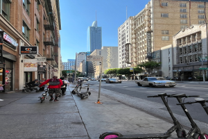 Eine Gruppe von Menschen, die auf Roller durch eine Stadtstraße fahren, mit hohen Gebäuden an beiden Seiten, einem geparkten Pick-up Truck, Straßenlaternen, Bäumen und Schildern.