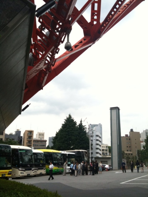 Menschen und Busse auf einer Straße mit Gebäuden, Bäumen und Himmel im Hintergrund und einer Brücke oben.