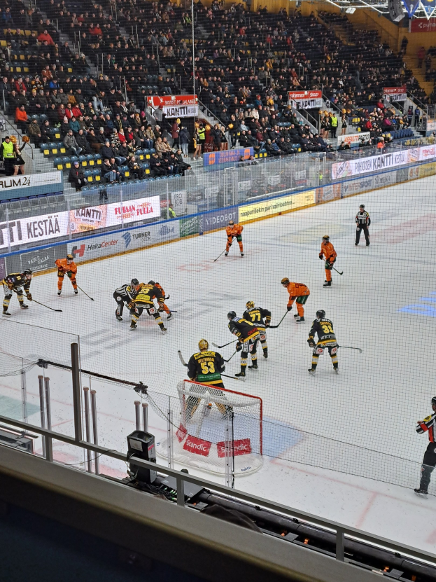 Eine Gruppe von Menschen, die Hockey auf einem Eisring spielen, umgeben von einem Zaun, mit Zuschauern im Hintergrund und beleuchteten Stadionlampen darüber.
