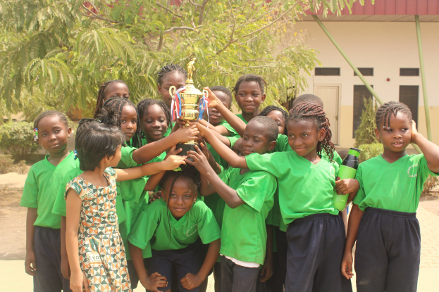 Eine Gruppe von Kindern, die eine Trophäe in einem Schulhof mit Bäumen, Pflanzen und einem Gebäude im Hintergrund halten.