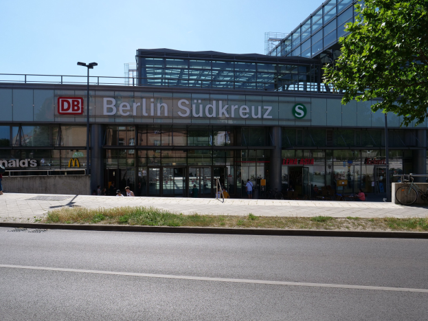 Großes gläsernes Berliner Südkreuz-Bahnhof mit Text an seiner Fassade, umgeben von Straßenlaternen, Straßenlaternen, Fahrzeugen, Fußgängern, Fahrrädern, Bäumen und einem klaren blauen Himmel.