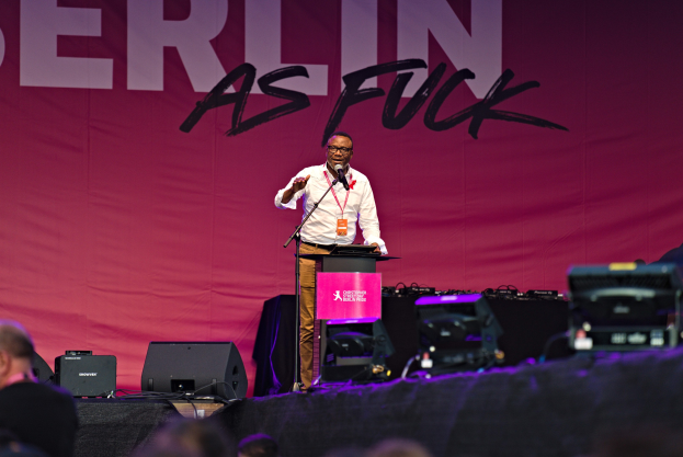 Ein Mann in einem weißen Hemd und braunen Hosen steht auf einer Bühne und hält ein Mikrofon in der Hand, hinter ihm eine pinkfarbene Fahne mit der Aufschrift 'Berlin as Fuck' und Lautsprecher auf der Bühne.