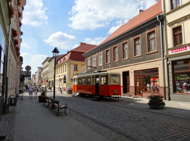 Eine rote und gelbe Straßenbahn fährt eine Kopfsteinpflasterstraße in Tallinns altem Stadtzentrum entlang, flankiert von Gebäuden mit Fenstern, Bänken und Laternen, mit Menschen auf den Gehwegen und einem bewölkten Himmel im Hintergrund.