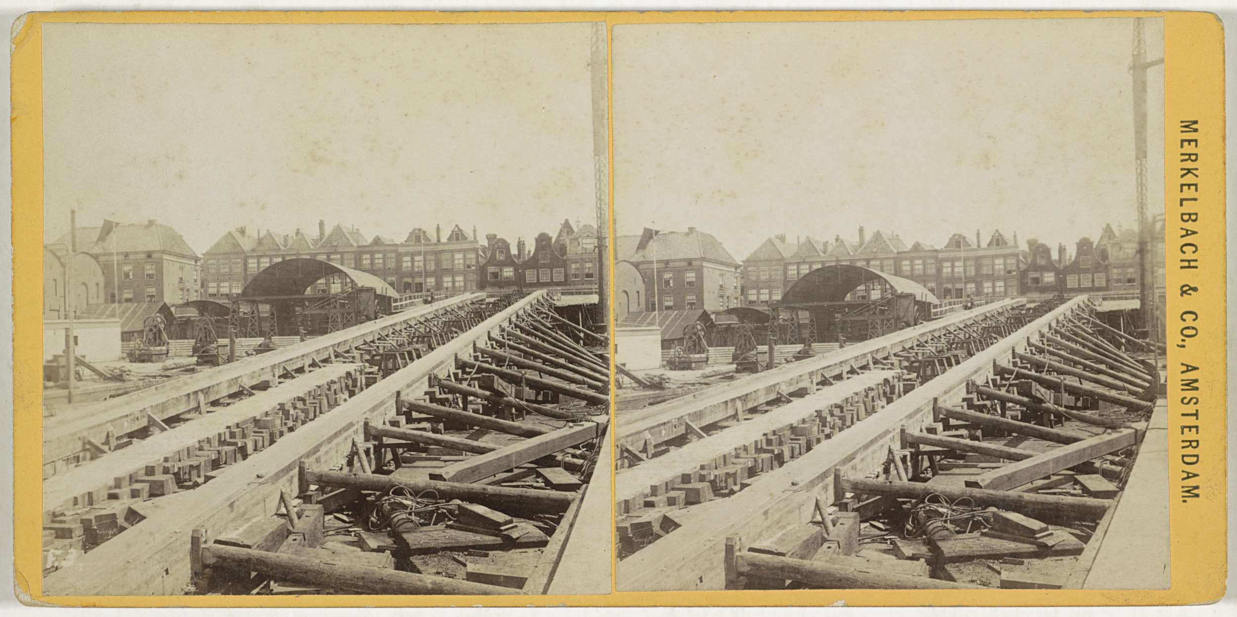 Ein altes Schwarz-Weiß-Foto einer Bahnschiene mit Gebäuden im Hintergrund, Text auf der rechten Seite mit 'Merkelbach & Co, Co. Amsterdam' und verstreuten Holzgegenständen im Vordergrund.