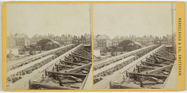 Ein altes Schwarz-Weiß-Foto einer Bahnschiene mit Gebäuden im Hintergrund, Text auf der rechten Seite mit 'Merkelbach & Co, Co. Amsterdam' und verstreuten Holzgegenständen im Vordergrund.