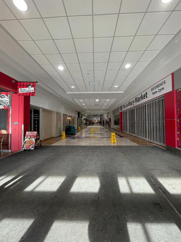 Ein hell erleuchtetes Einkaufszentrum-Innere mit einem Stuhl und einer Tafel, die links " Sydbury Market - Vorher und Nachher" trägt, und verstreuten Gegenständen auf der rechten Seite, mit dem Boden darunter.