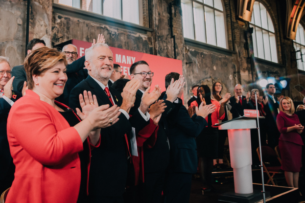 Eine Gruppe von Menschen, die vor einem Publikum jubelnd klatschen, mit einem Podium, einem Mikrofon und einer Tafel mit Text auf der rechten Seite und Stühlen, einem Banner, einer Wand, Fenstern und Lichtern im Hintergrund.