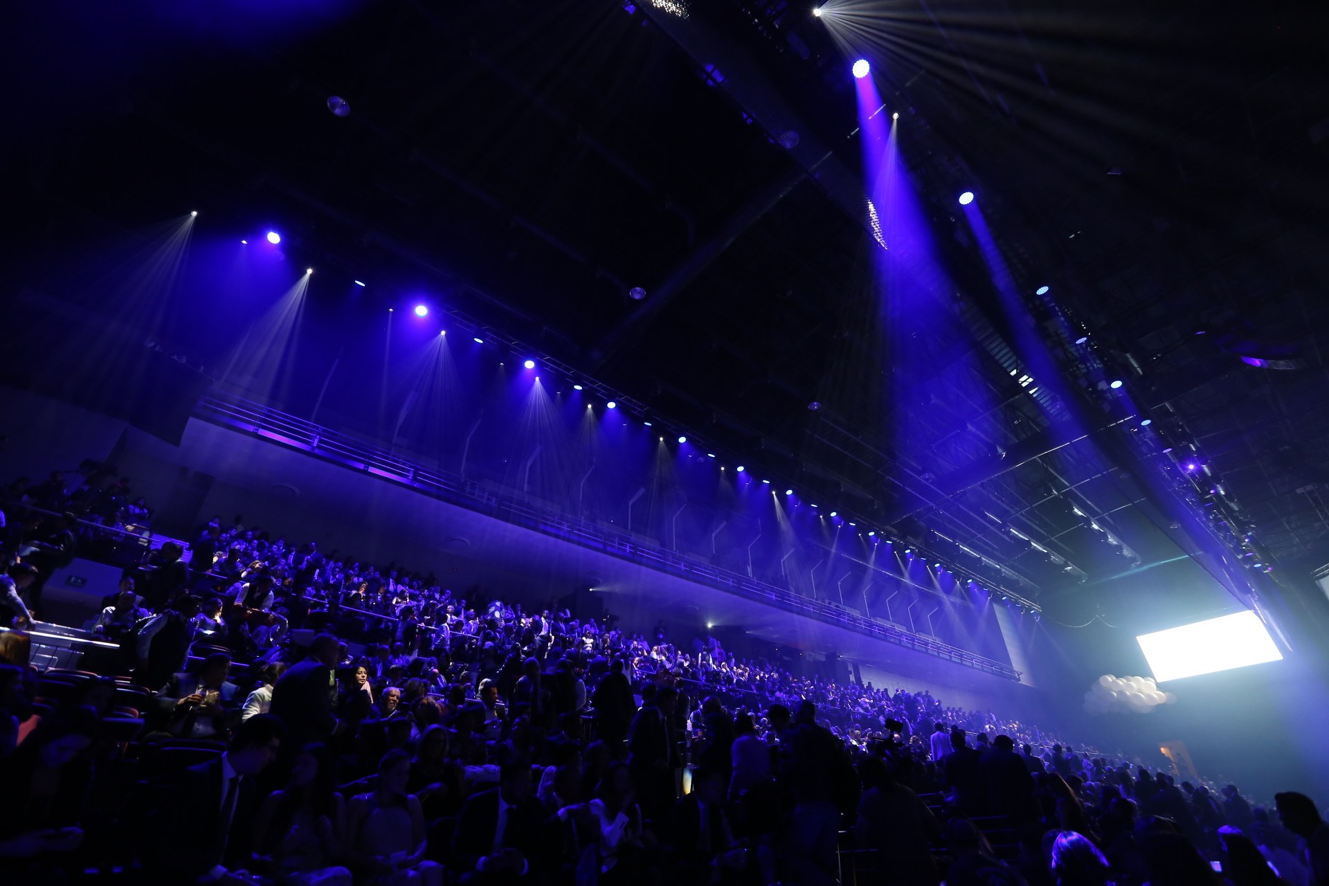 Große Menschenmenge in einem abgedunkelten Konzertsaal gegenüber einer Bühne mit deckenmontierten Lichtern und einem Bildschirm auf der rechten Seite.