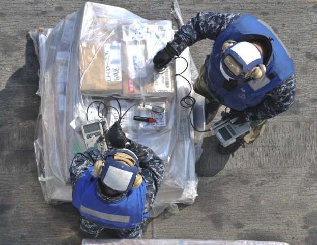 Zwei Männer in blauen Uniformen und Helmen arbeiten an einem großen Drohnen, halten verschiedene Gegenstände, während eine Plastikabdeckung auf dem Boden liegt.
