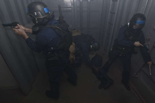 Eine Gruppe von Polizisten in Einsatzausrüstung, einer hält eine Waffe, steht neben einem Metallcontainer und einer Leiter in einem begrenzten Raum.