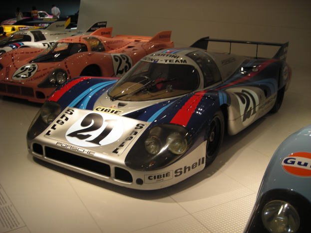 Ein Porsche 917K Rennwagen in einem Museum ausgestellt, umgeben von anderen Sportwagen, mit Menschen in der Nähe und einer Wand im Hintergrund.
