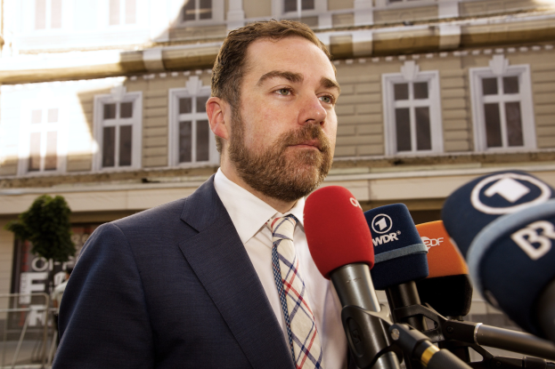 Mann im Anzug und Krawatte spricht vor der Kamera vor einem Gebäude, wahrscheinlich über eine dreijährige Haftstrafe.