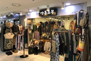 Kleidungsgeschäftsinnere mit Ständern voller Kleider, Hüten, Taschen und Schuhen, Deckenlampen und Informationsschildern.