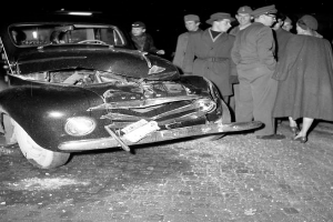 Eine schwarz-weiße Szene eines beschädigten Autos am Straßenrand, umgeben von einer Gruppe besorgter Menschen.