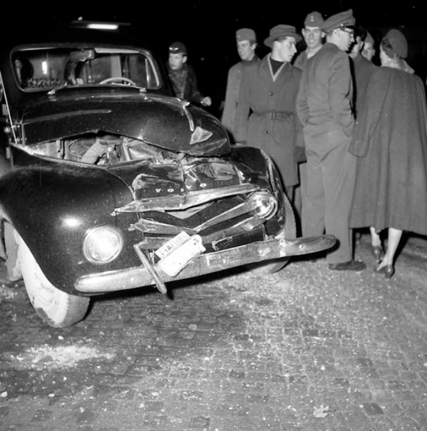 Eine schwarz-weiße Szene eines beschädigten Autos am Straßenrand, umgeben von einer Gruppe besorgter Menschen.