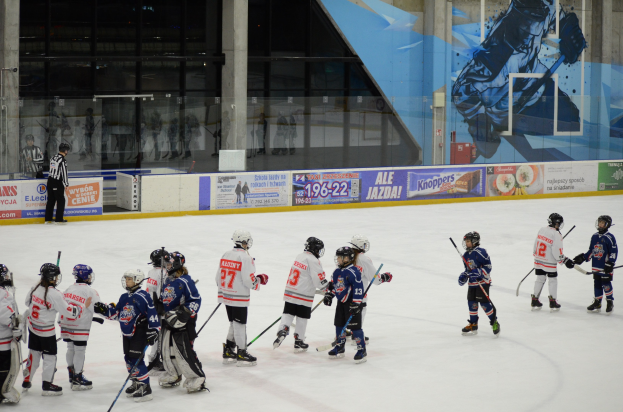 Junge Menschen in Helmen und Hockey-Ausrüstung spielen Eis Hockey auf einer Indoor-Eisbahn mit Bannern, Säulen und Glaswänden im Hintergrund.
