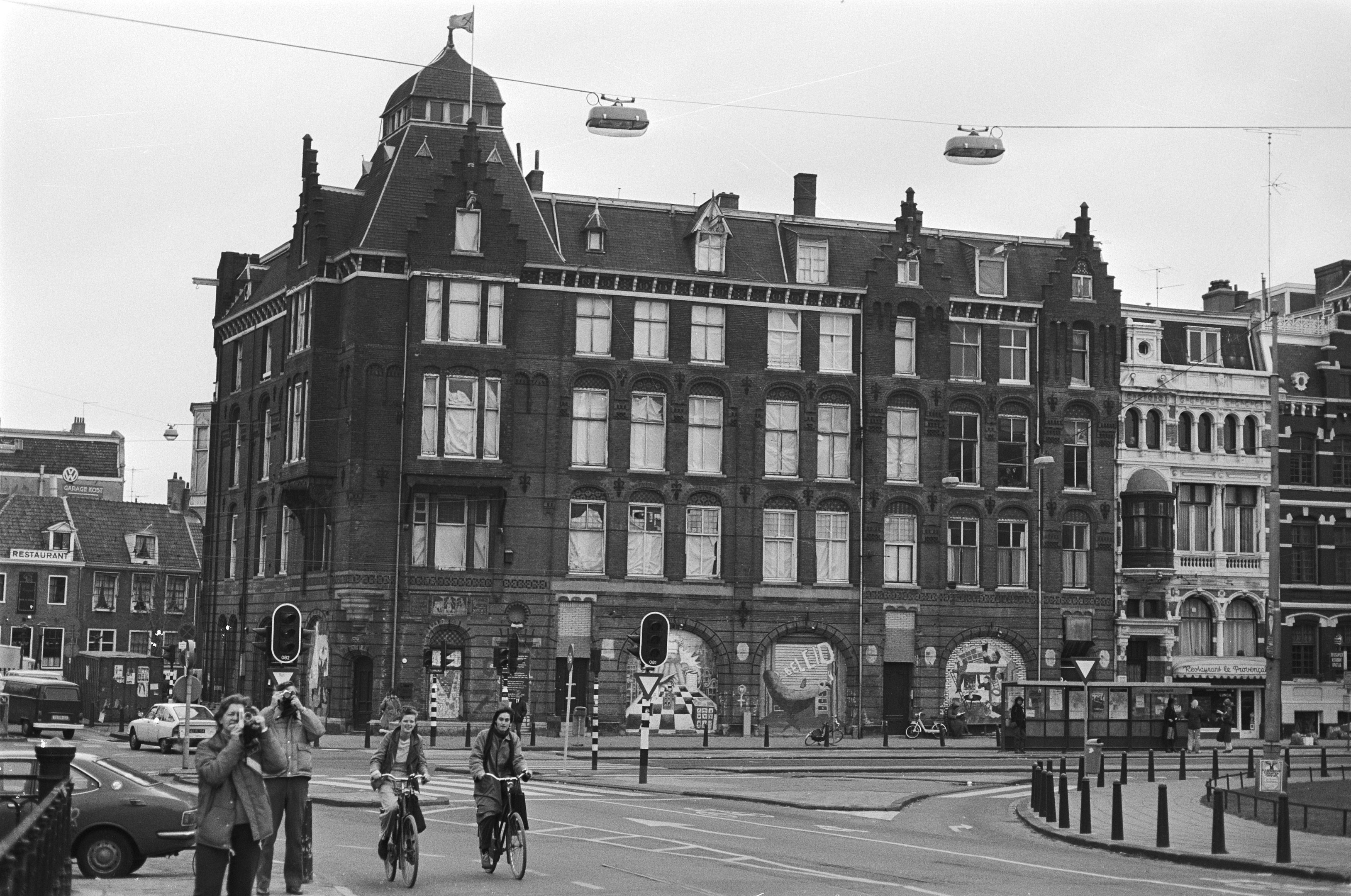 Schwarzes und weißes Foto von Menschen, die auf der Straße mit Gebäuden, Straßeninfrastruktur, Kraftfahrzeugen, Fußgängern und Himmel Fahrräder fahren.