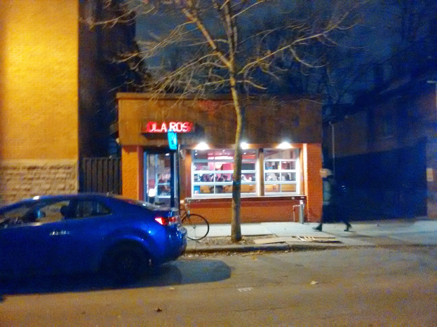 Ein Auto fährt nachts auf einer Straße, mit einer Person, die auf einem Fahrrad auf einem Gehweg neben ihm fährt, einem Baum, einem Geschäft und einigen Gebäuden im Hintergrund.