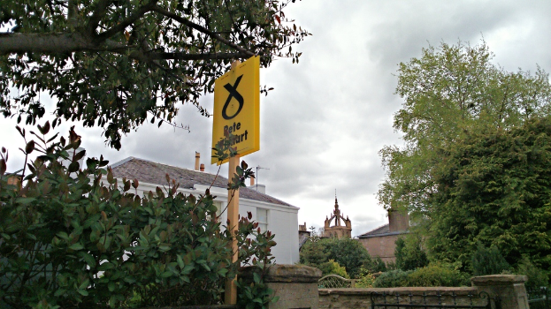 Ein gelbes Warnschild auf dem Boden vor einem weißen Schattendach, mit einer braunen Kirche und Bäumen im Hintergrund und einer kleinen Begrenzungsmauer vorne.