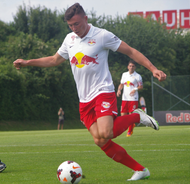 Ein Mann in einem Fußballtrikot schießt einen Ball auf einem grünen Feld mit Bäumen und einem klaren blauen Himmel im Hintergrund, mit einem Schild mit der Aufschrift "RB Leipzig" im Hintergrund.