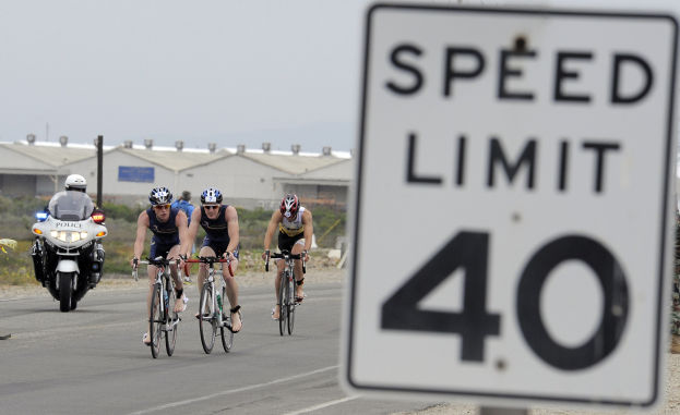 Gruppe von Radfahrern mit Helmen auf einer Straße neben einem Geschwindigkeitsbegrenzungsschild mit Gebäuden, Masten, Pflanzen, Bäumen und einem klaren blauen Himmel im Hintergrund.