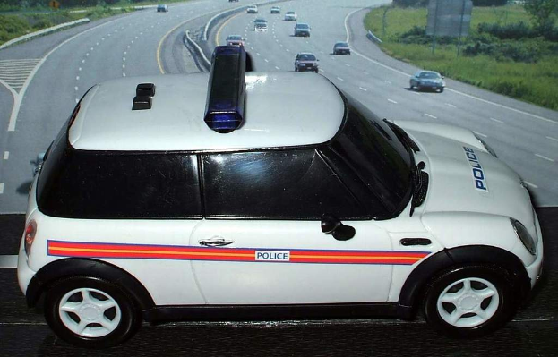 Ein weißer Polizeiwagen steht im Vordergrund, hinter ihm fahren mehrere Autos auf der Straße, umgeben von Gras und Bäumen auf beiden Seiten.