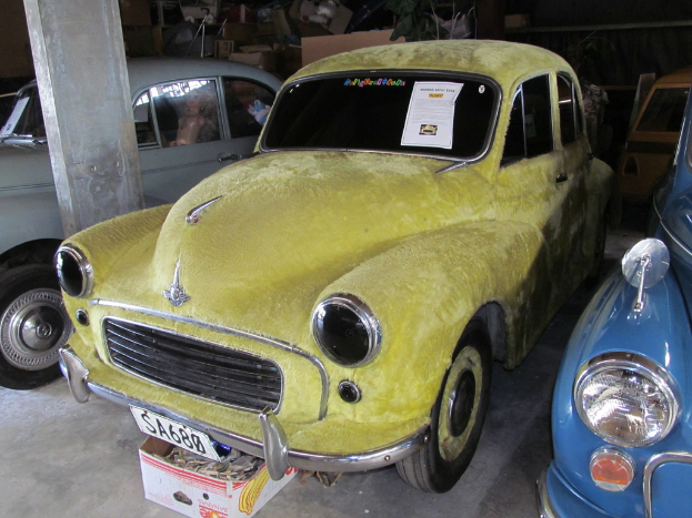 Mehrere Oldtimer sind vor einer Wand mit verschiedenen Accessoires geparkt, wobei links eine Säule zu sehen ist.