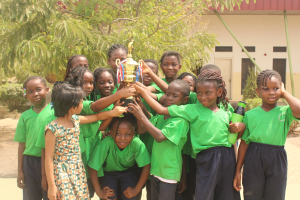 Eine Gruppe von Kindern, die zusammenstehen und jeweils einen Pokal hochhalten, mit Bäumen, Pflanzen und einem Gebäude im Hintergrund.