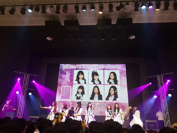 Eine K-Pop-Band, BK48, performs auf einer hell erleuchteten Bühne mit zwei Mitgliedern, die ein Mikrofon halten und ein Instrument spielen, während eine Menge darunter das Konzert aufnimmt.