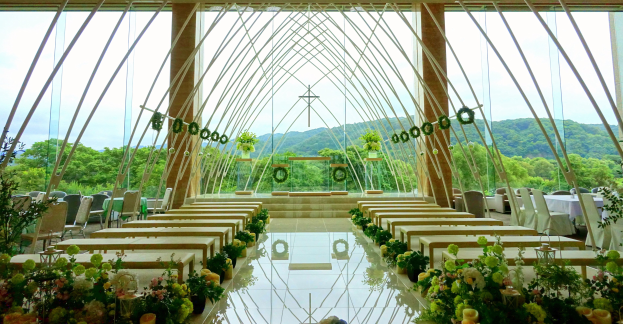 Eine Trauungszeremonie in einem großen Saal mit halbrunder Bestuhlung, Blumenschmuck und einem Blick auf Bäume, Hügel und einen klaren Himmel im Hintergrund.