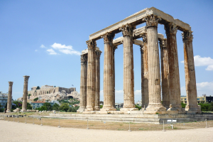 Alter Tempel von Olympian Zeus in Athen, Griechenland, mit hohen korinthischen Säulen, umgeben von Gebäuden, Bäumen und felsigem Gelände, mit einer Festung in der Ferne, einem bewölkten Himmel.