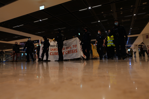 Eine Gruppe maskierter Polizisten in einem Flughafen, die ein Banner halten, umgeben von Geländern und Deckenleuchten, mit Texttafeln im Hintergrund.