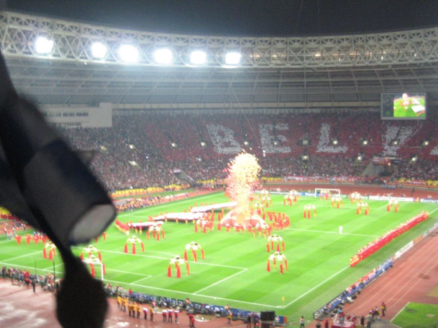 Ein Stadion mit Flutlicht, Eisengittern, Zuschauern, stehenden Personen, Werbetafeln, Anzeigetafeln, Sicherheitskameras und dekorativen Fahnen.