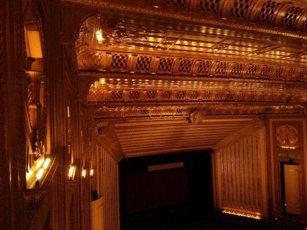 Ein prunkvoller Theatersaal mit dunklem Hintergrund, beleuchtet von Lichtern links und rechts, mit einer kunstvoll gestalteten Decke.