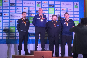 Fünf medaillenreiche Männer auf einem Podium mit einem Fotografen, der den Moment einfängt, und einem Banner mit der Aufschrift "DDBU-Weltmeisterschaft 2019" im Hintergrund.